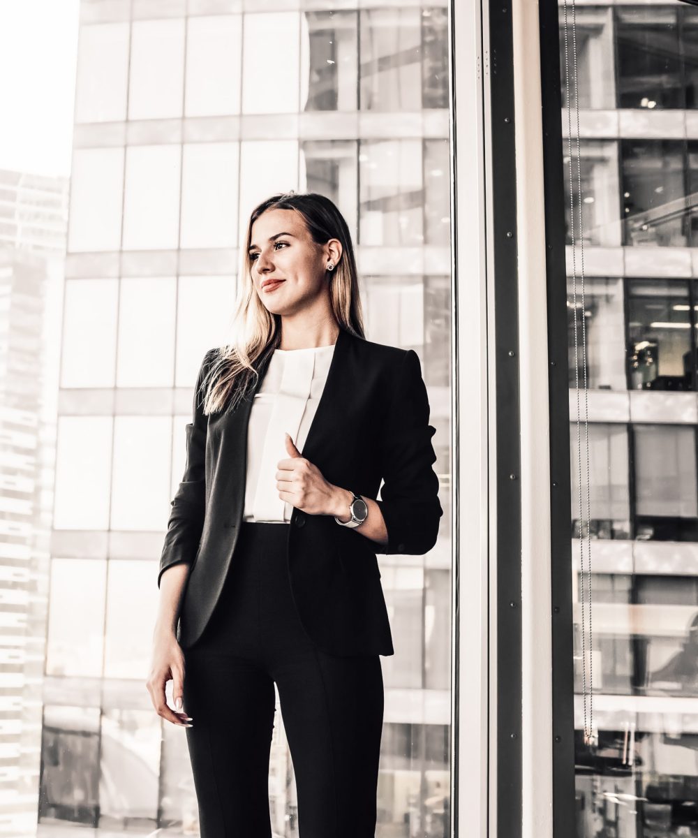 Cheerful blonde female manager in formal outfit enjoying job and successful career in business, smiling beautiful woman boss dressed in elegant suit standing near panoramic windows in office