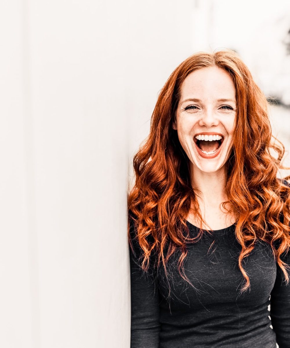 Cute young woman with a lovely sense of humour standing leaning against a white exterior wall with copy space in an urban street laughing at the camera